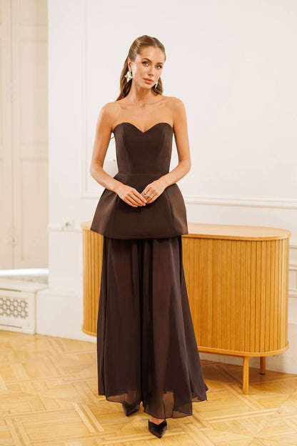 A woman models a dark brown strapless gown with a sweetheart neckline, structured peplum waist, and a flowing chiffon skirt, standing in a bright room with a wooden cabinet.