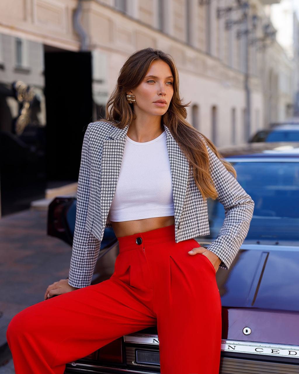 Woman in a checkered blazer, white top, and red pants sitting on a car in an urban setting.