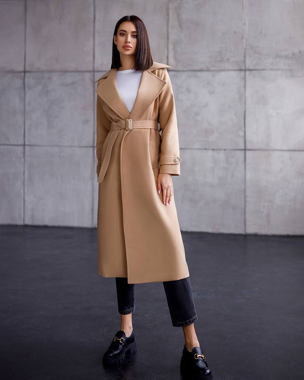 Woman wearing a beige trench coat against a gray wall.