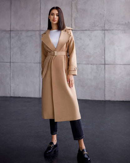 Woman wearing a beige trench coat against a gray wall.