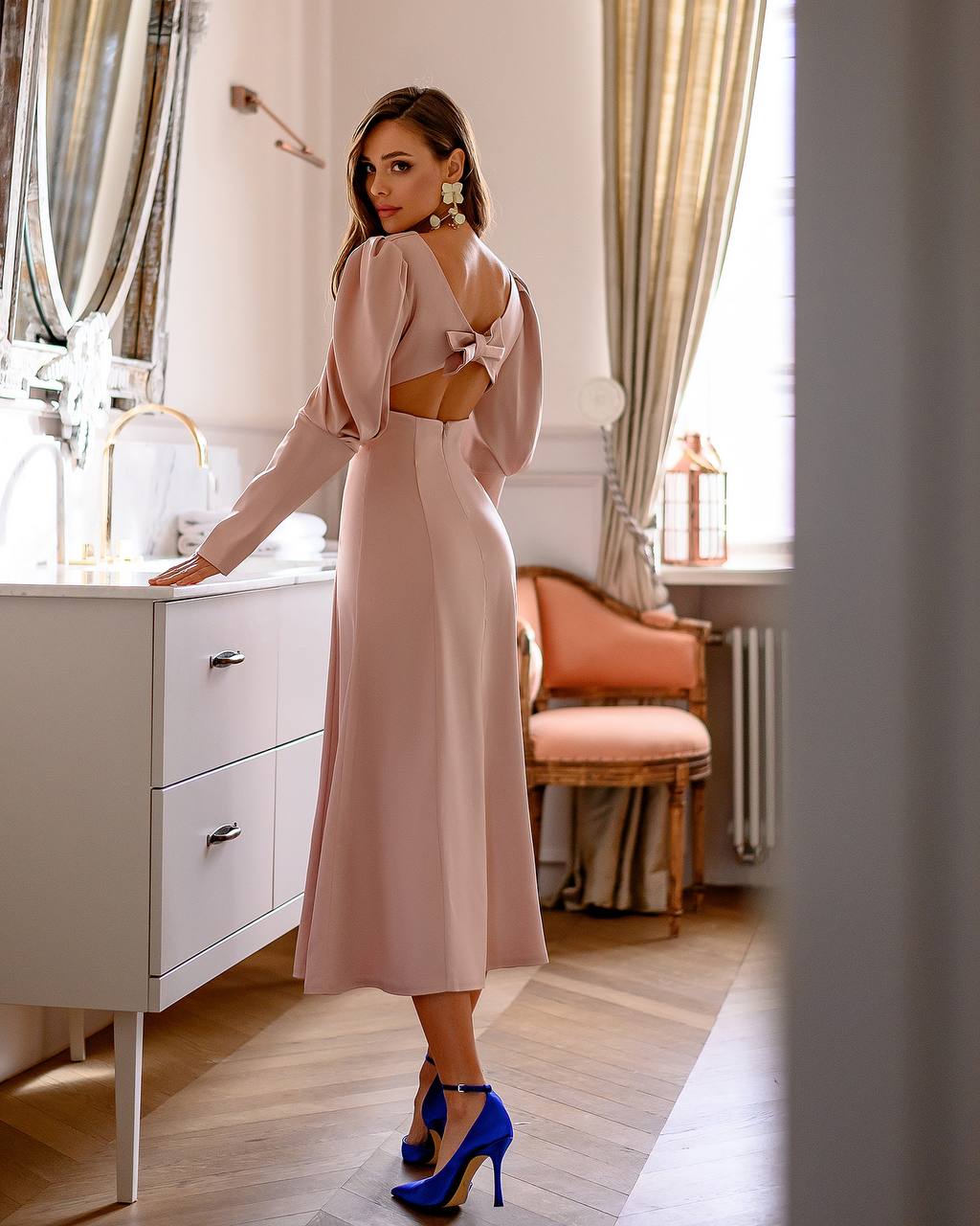 Woman in a pink dress standing in a stylish room with a mirror and chair.