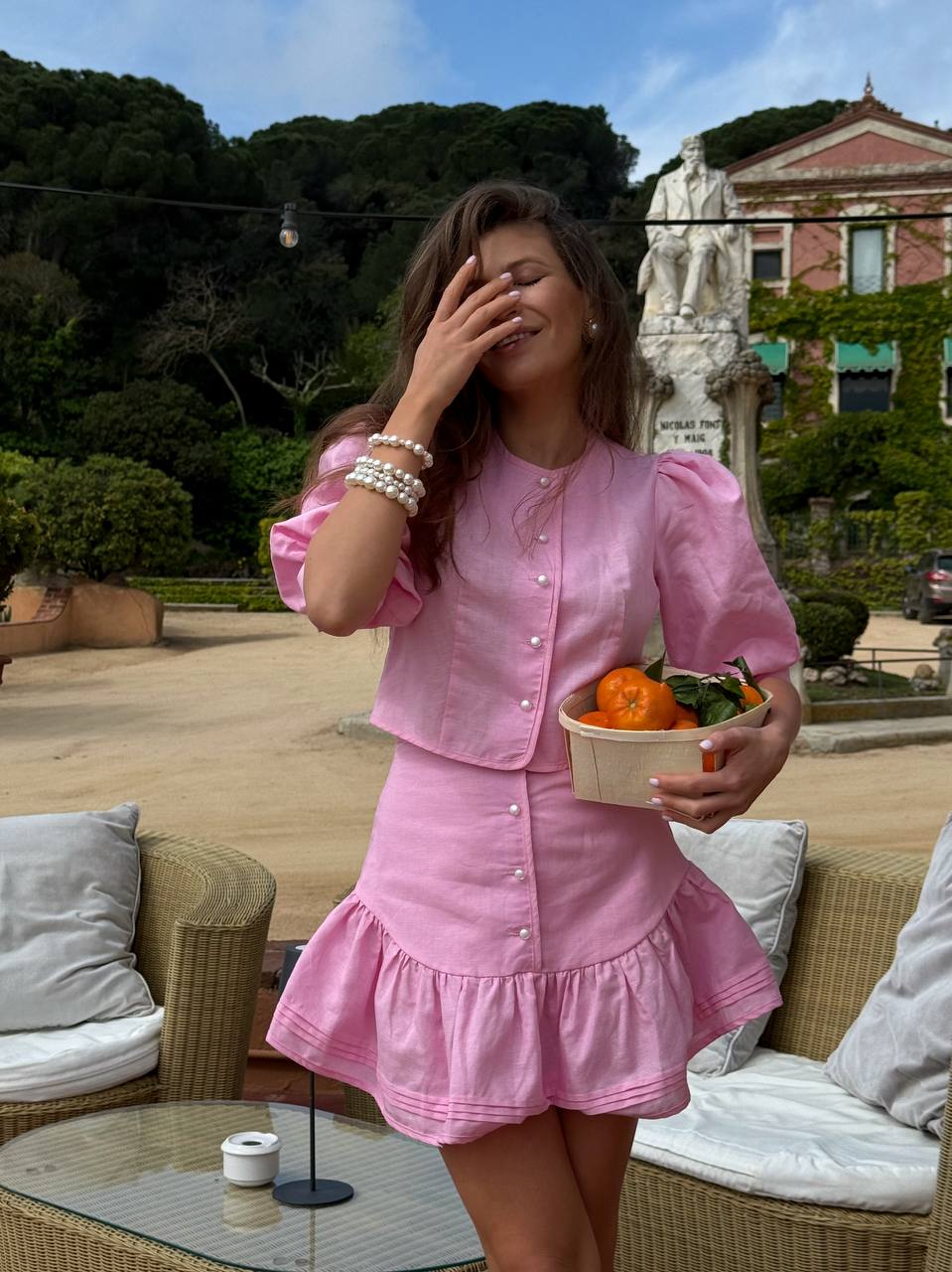 Woman in a pink outfit holding a bowl of food outdoors with greenery and a building in the background.