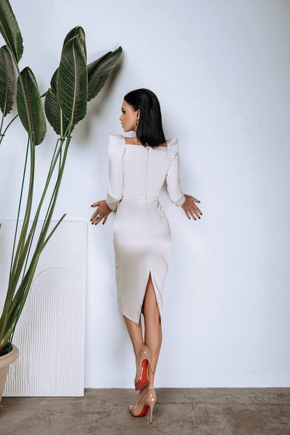 Woman in a white dress standing next to a large plant against a light gray wall.