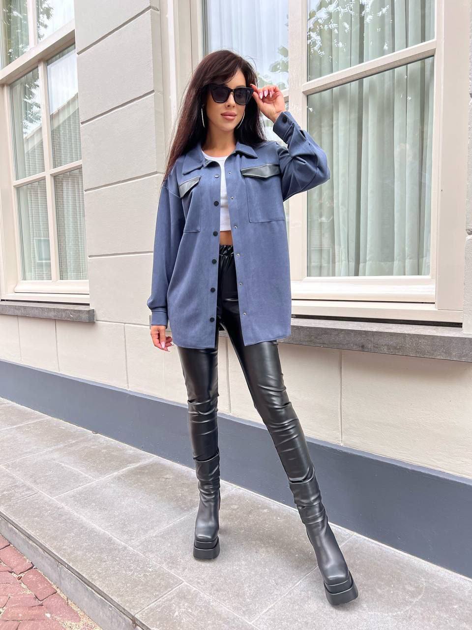 Woman wearing a blue denim jacket, white top, black leather pants, and black boots standing outside a building.