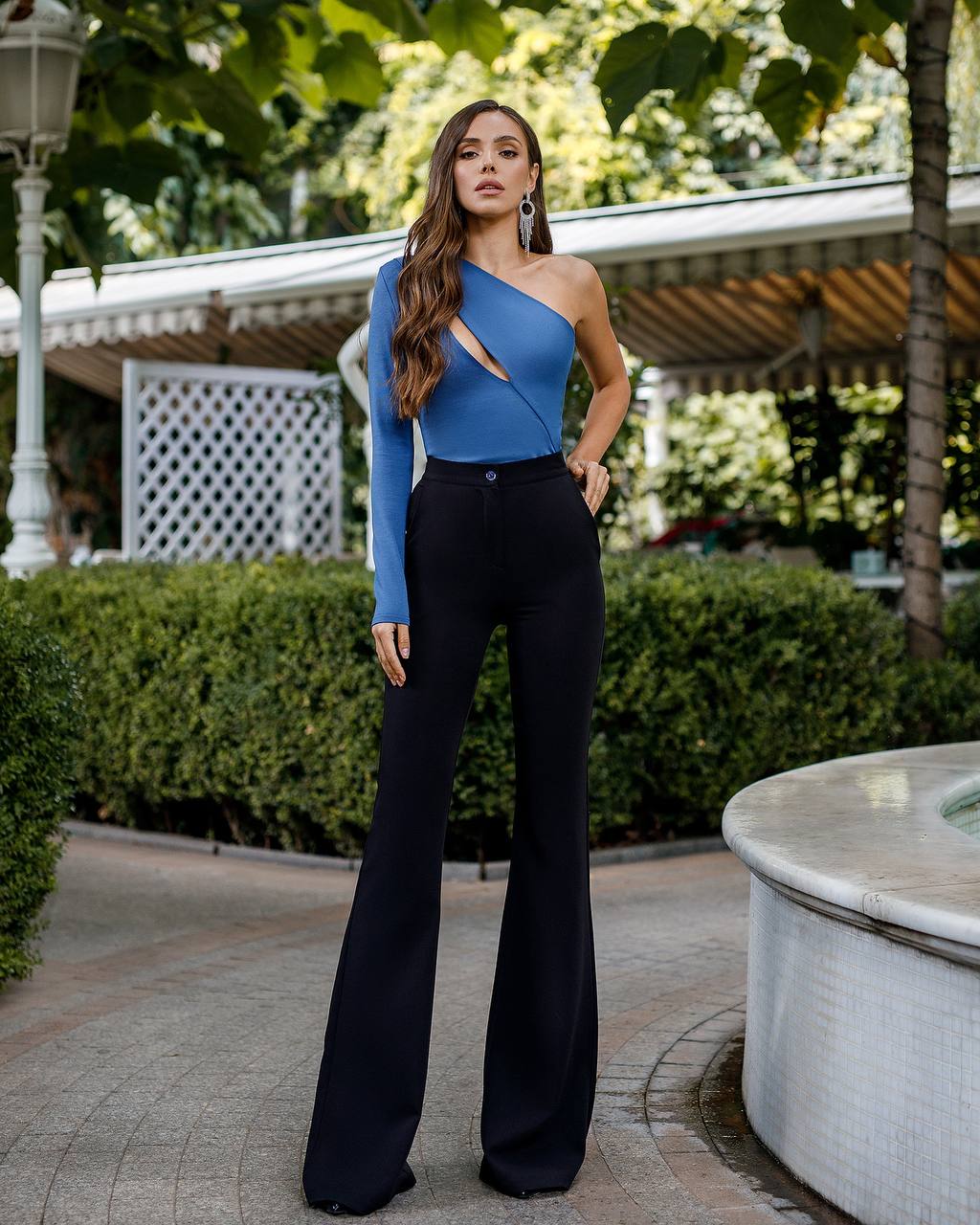 Woman in a blue top and black pants standing outdoors with greenery in the background