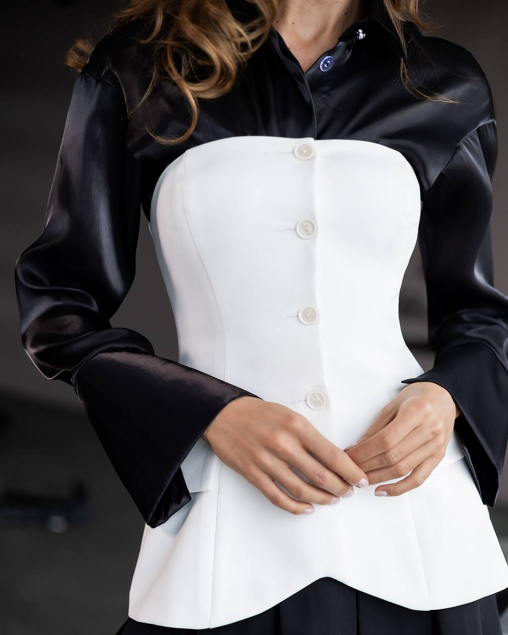A close-up of a model wearing a structured white corset-style top with a black satin long-sleeve collar and cuffs, fastened with white buttons.