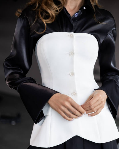 A close-up of a model wearing a structured white corset-style top with a black satin long-sleeve collar and cuffs, fastened with white buttons.
