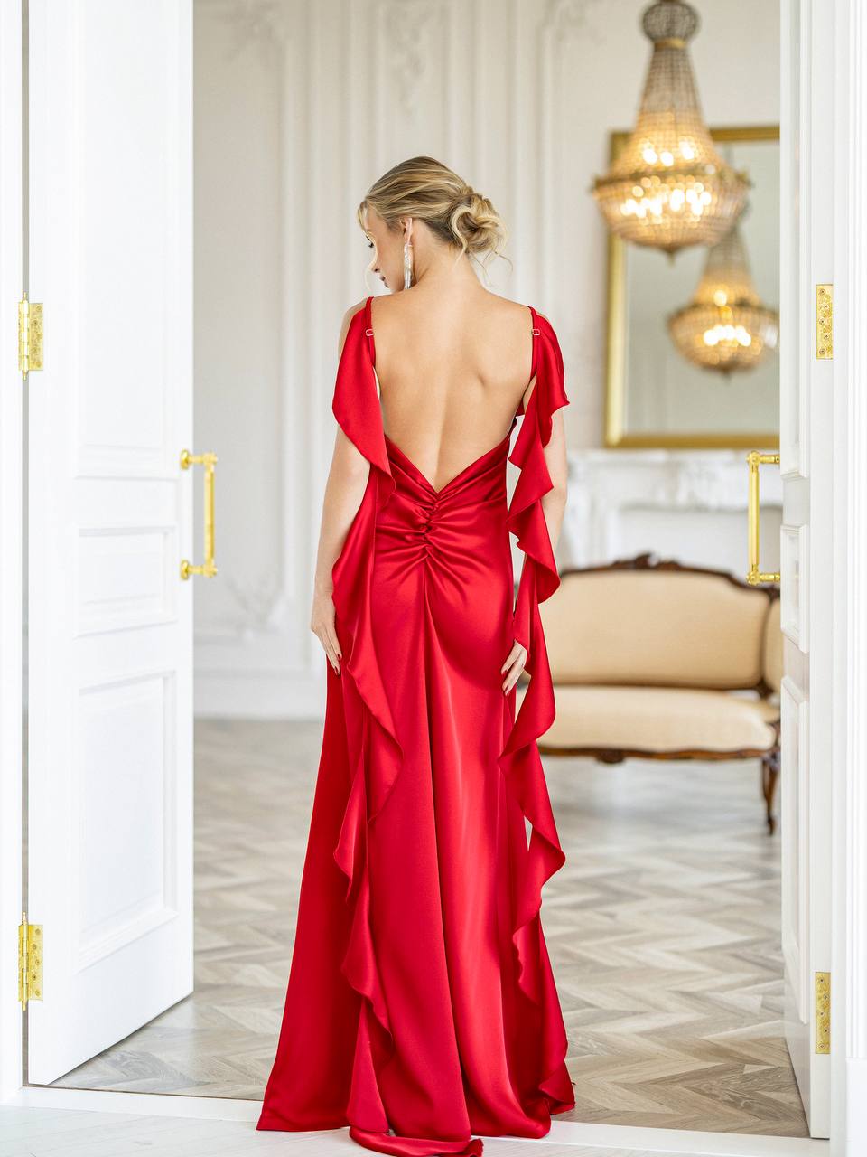 A woman wearing a bright red satin evening gown with a dramatic open back and layered ruffle details, standing in a doorway of a classic white room with gold handles and chandeliers.