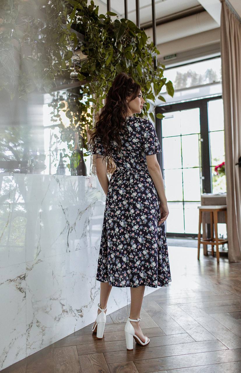 Woman in a floral dress standing in a modern interior setting with plants and marble wall.