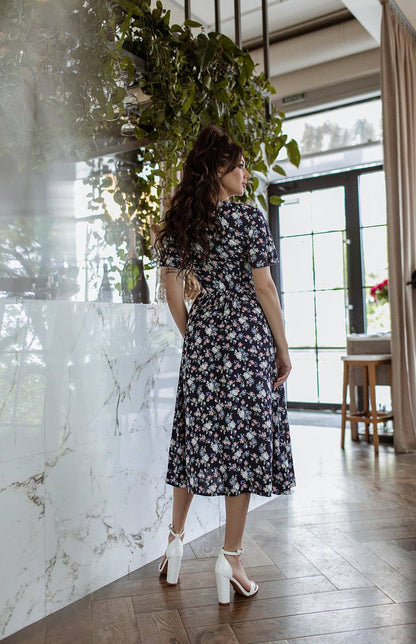 Woman in a floral dress standing in a modern interior setting with plants and marble wall.
