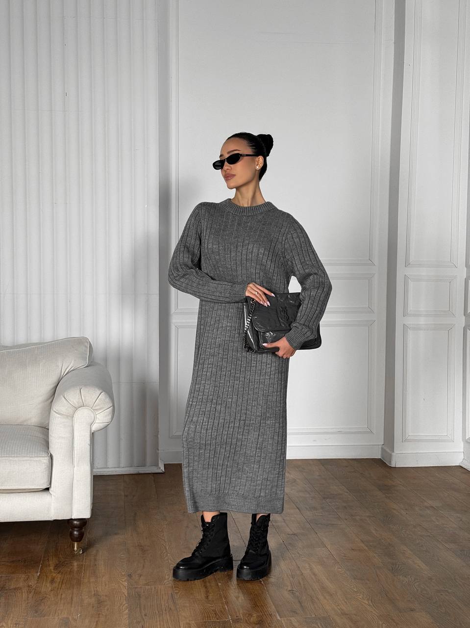 Woman in a gray knit dress holding a black clutch in a room with white walls and a gray sofa.