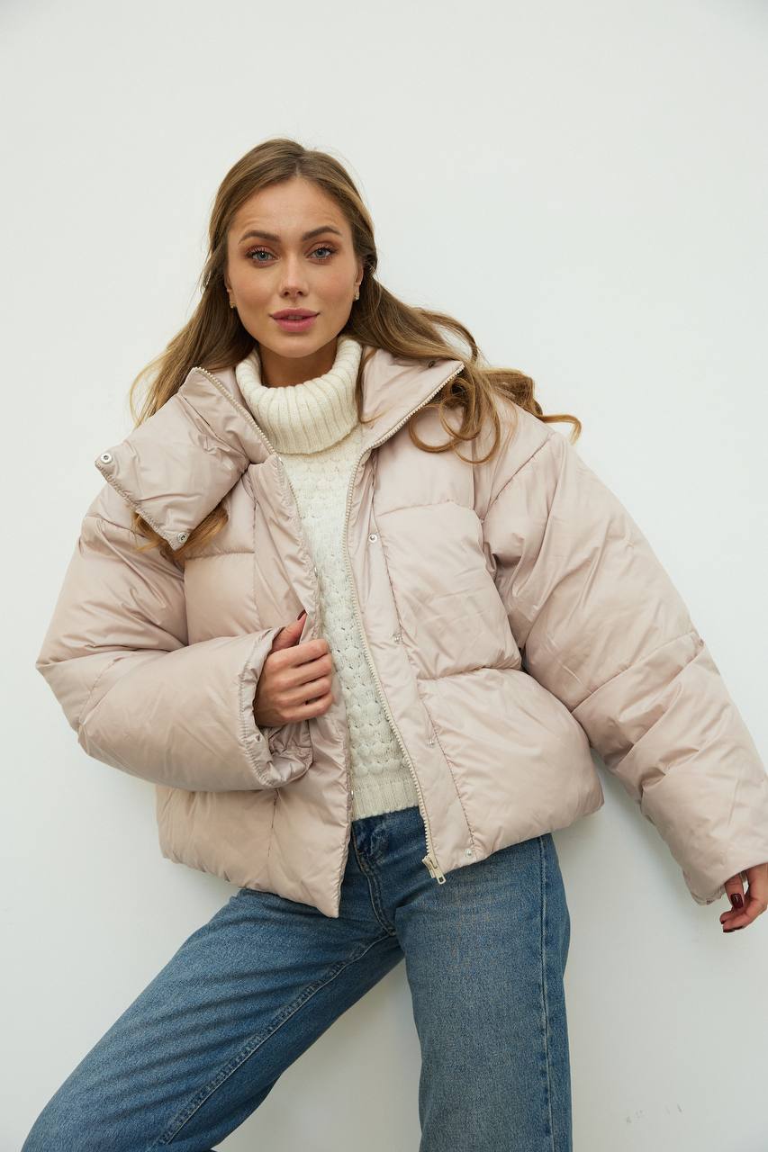 Woman wearing a beige puffer jacket over a white sweater and blue jeans against a white background