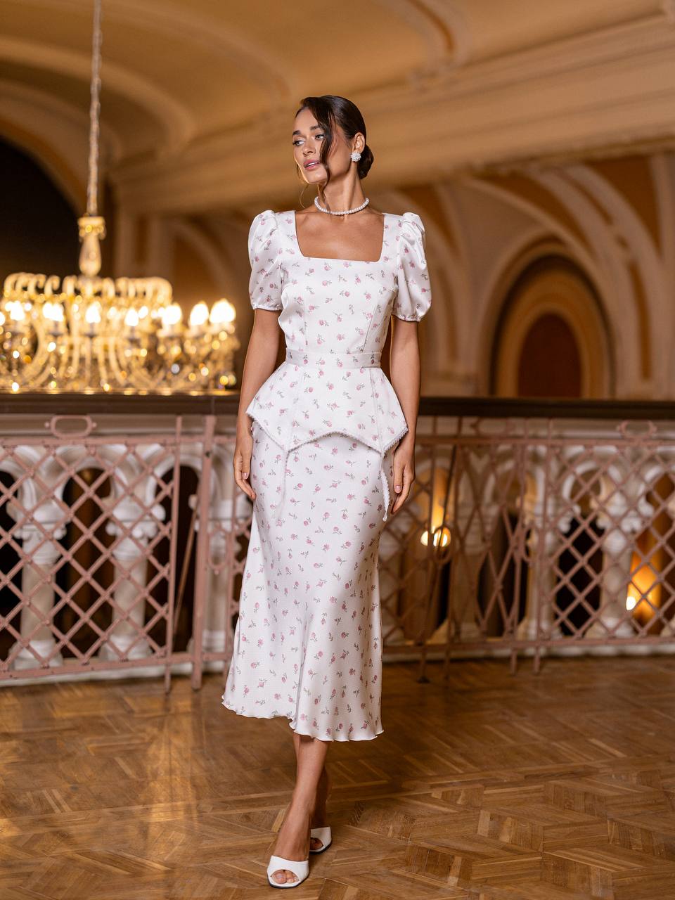 Woman in a white dress standing in an elegant indoor setting with chandeliers.