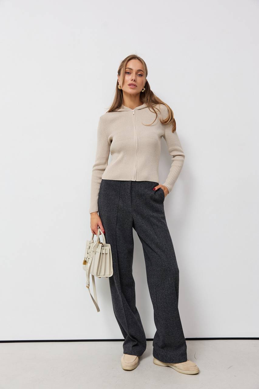 Woman wearing a beige sweater and gray pants against a white background