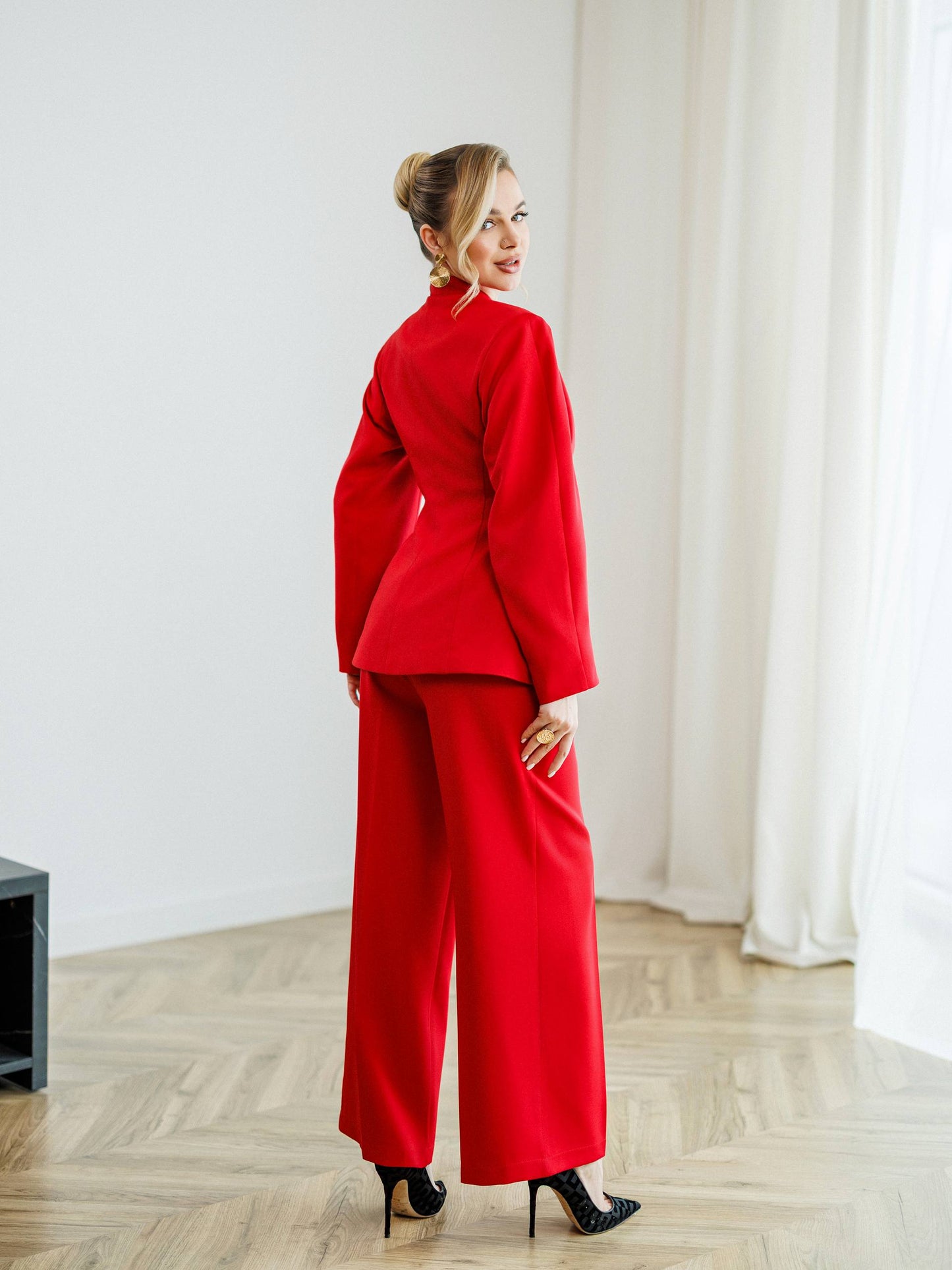 Woman in a red outfit standing in a room with white curtains and wooden floor.