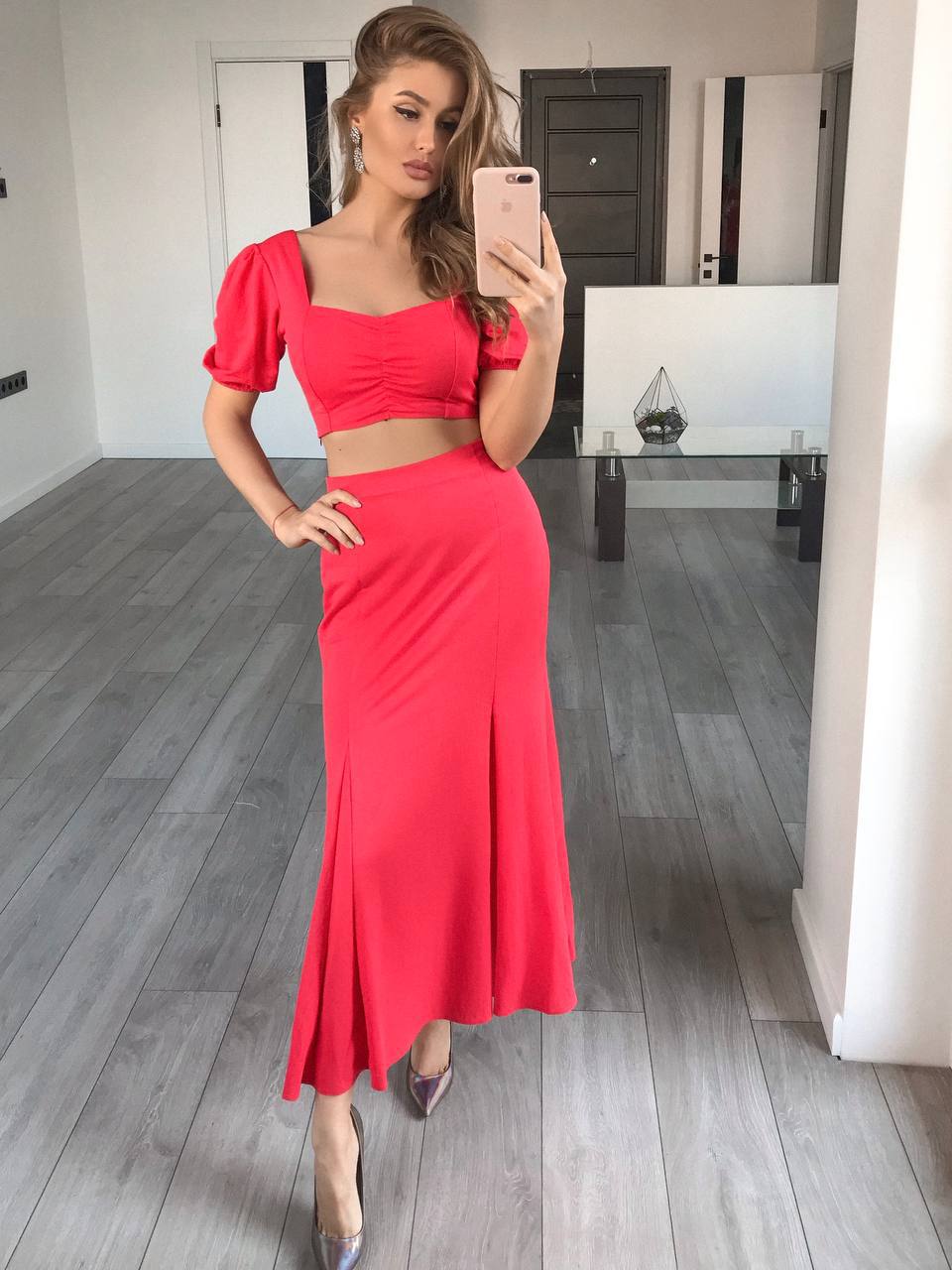 A woman takes a mirror selfie in a modern room with gray wood floors, wearing a bright coral-red two-piece dress with a square-neck crop top and a flared maxi skirt.