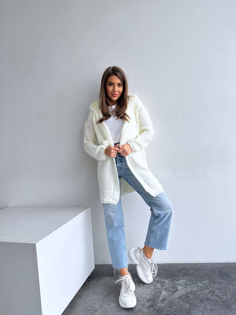 Woman wearing a white sweater, white shirt, blue jeans, and white sneakers against a gray wall.