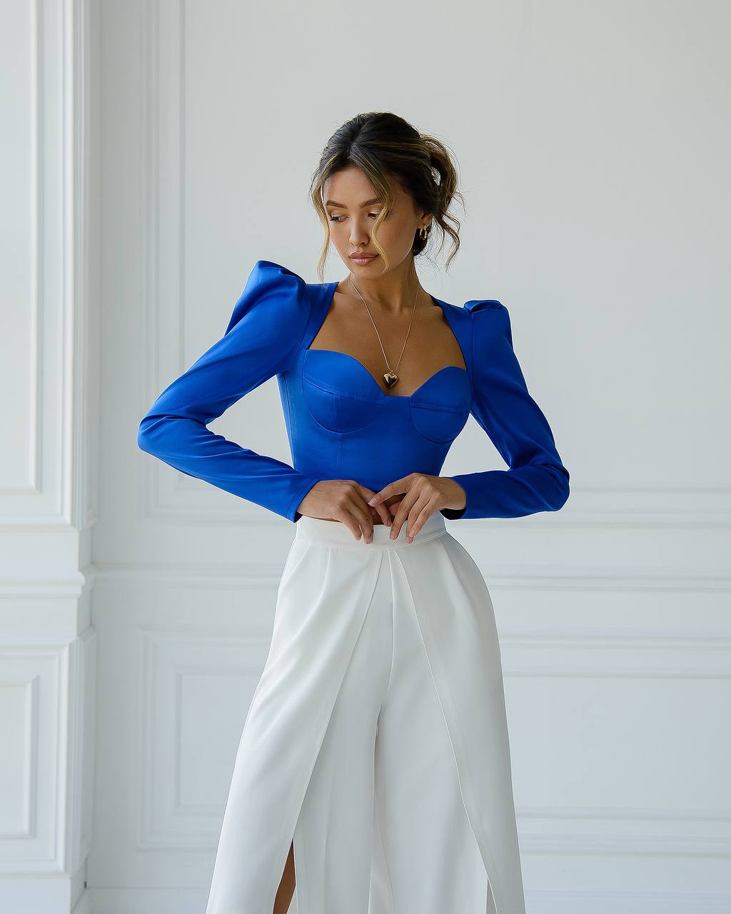 Woman wearing a blue top and white pants against a white wall