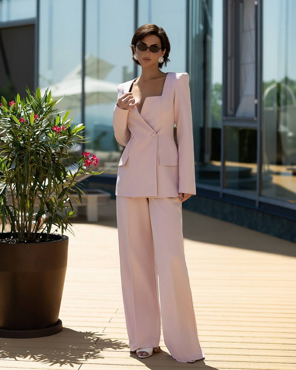A woman stands outdoors on a wooden deck wearing a light blush pink tailored suit with a structured blazer and wide-leg trousers. She is wearing dark sunglasses and white heeled sandals, with a large potted plant and modern glass building in the background.