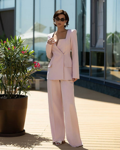 A woman stands outdoors on a wooden deck wearing a light blush pink tailored suit with a structured blazer and wide-leg trousers. She is wearing dark sunglasses and white heeled sandals, with a large potted plant and modern glass building in the background.