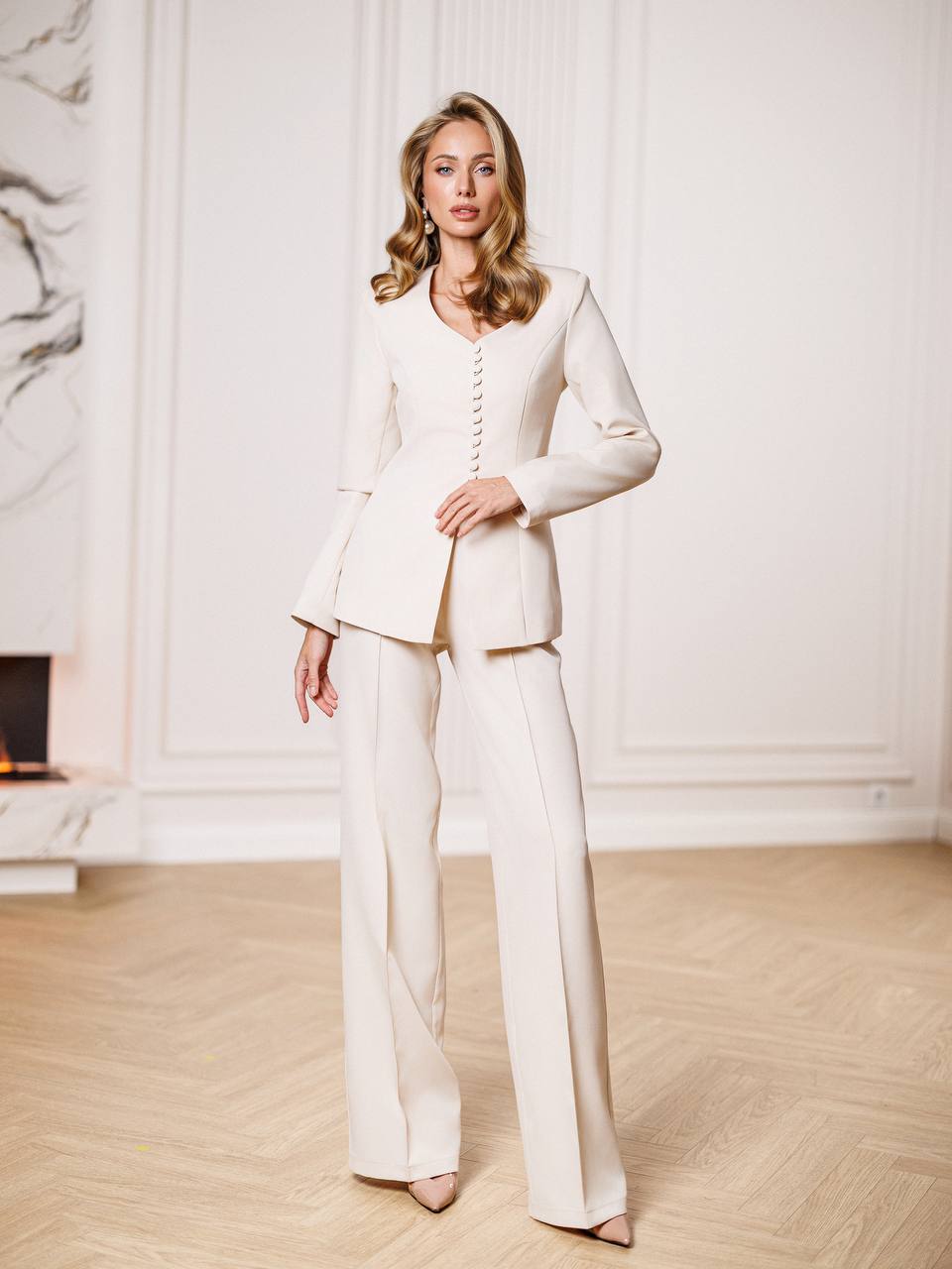 Woman wearing a white suit standing in a room with a fireplace and white walls.