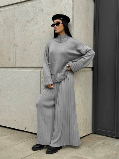 Person wearing a gray outfit with a pleated skirt against a neutral wall.