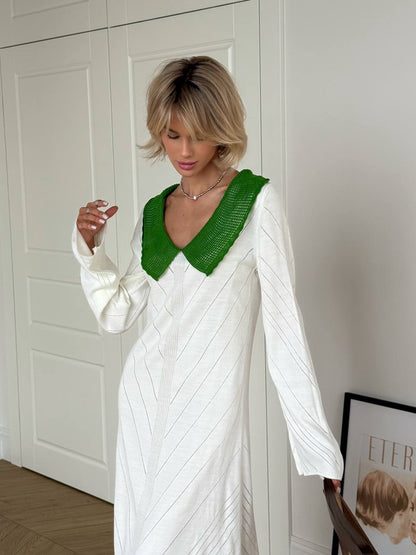 Woman wearing a white dress with a green neckline against a white wall.
