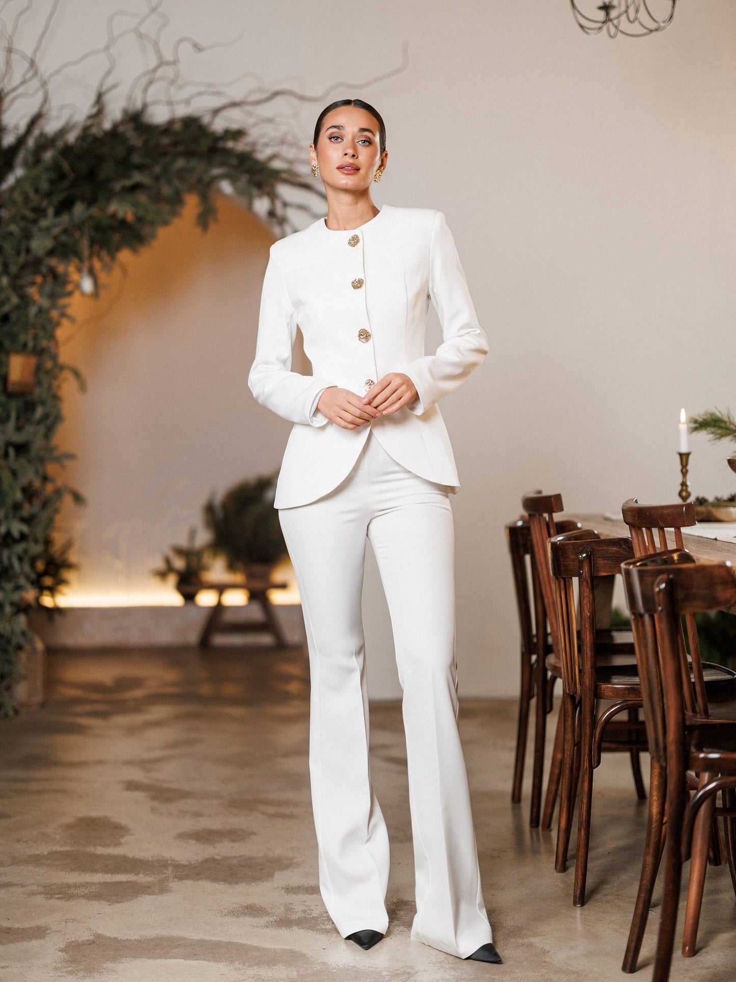 Woman in a white suit standing in an elegant indoor setting with wooden chairs and plants.