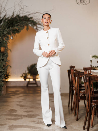 Woman in a white suit standing in an elegant indoor setting with wooden chairs and plants.