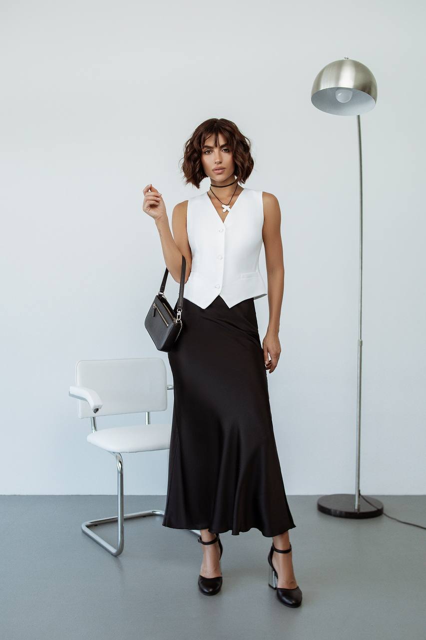 Woman in a white sleeveless top and black skirt standing in a minimalistic room with a chair and lamp.