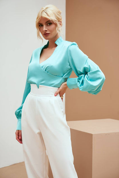 Woman wearing a turquoise blouse with puffed sleeves and white pants against a neutral background