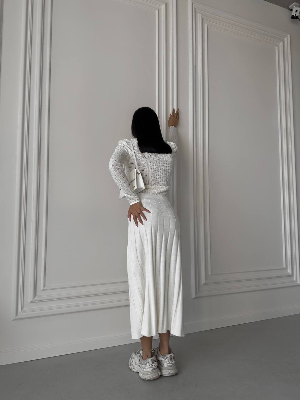 Person wearing a white outfit standing against a gray paneled wall.