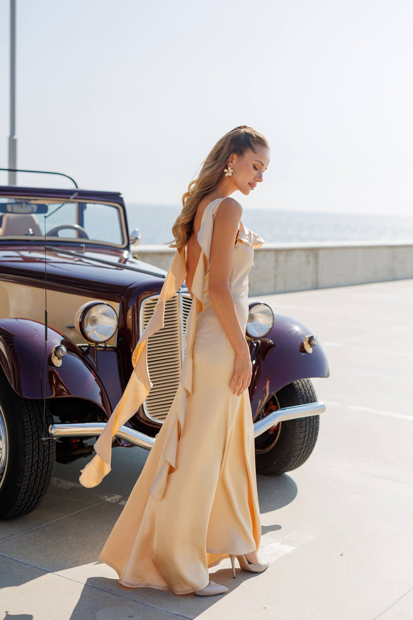 Woman in a long beige dress standing next to an old-fashioned car with a clear sky background