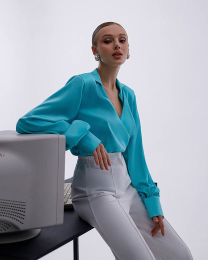 Person wearing a turquoise blouse and white pants sitting at a desk with a computer monitor.