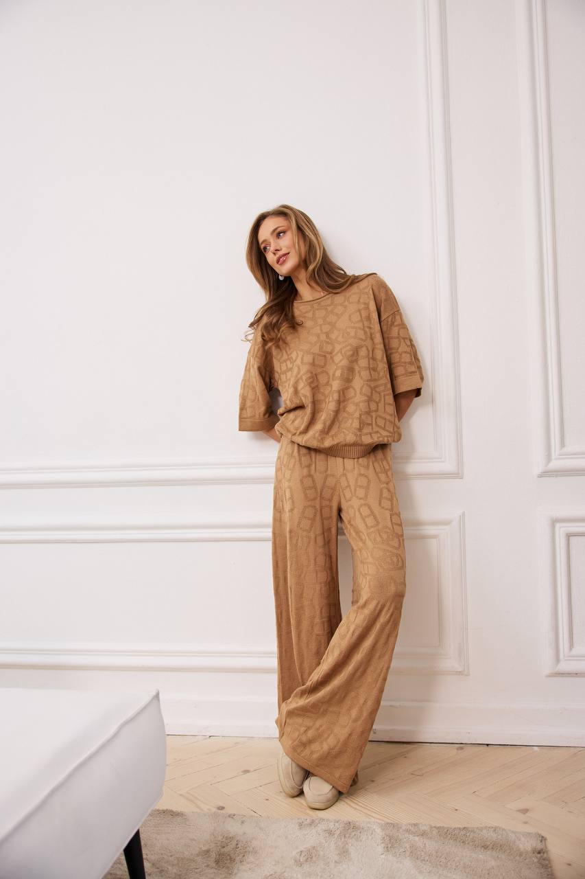 Woman wearing a beige textured outfit standing against a white paneled wall.