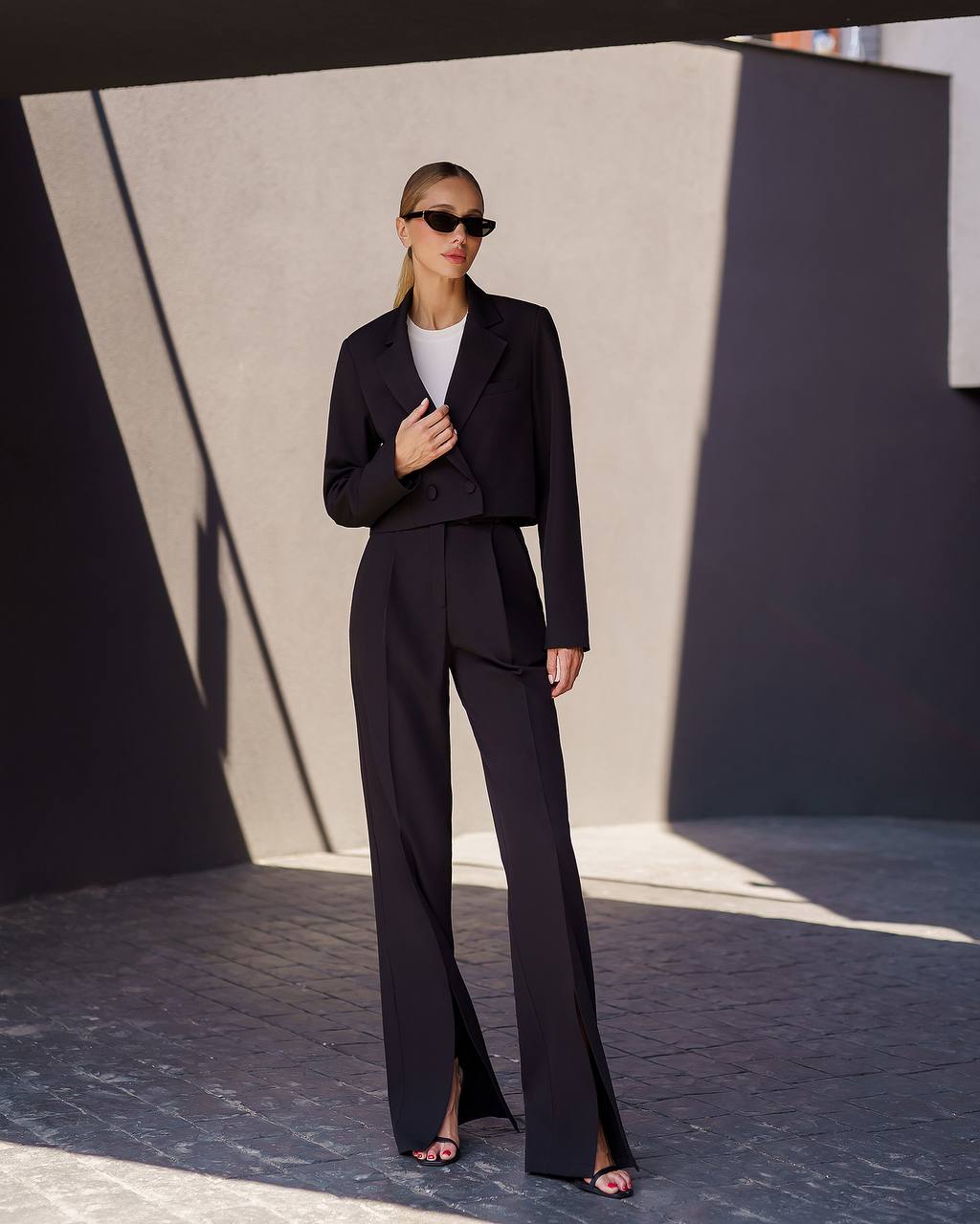 Woman in a black suit standing against a textured wall with geometric shadows.