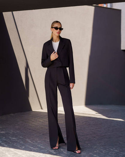 Woman in a black suit standing against a textured wall with geometric shadows.