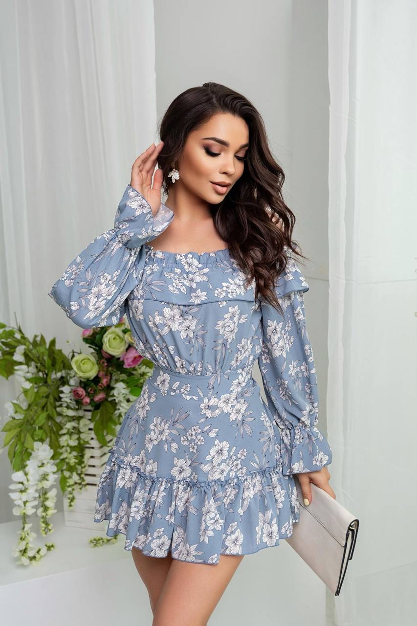 Woman wearing a blue floral dress holding a clutch against a white curtain background