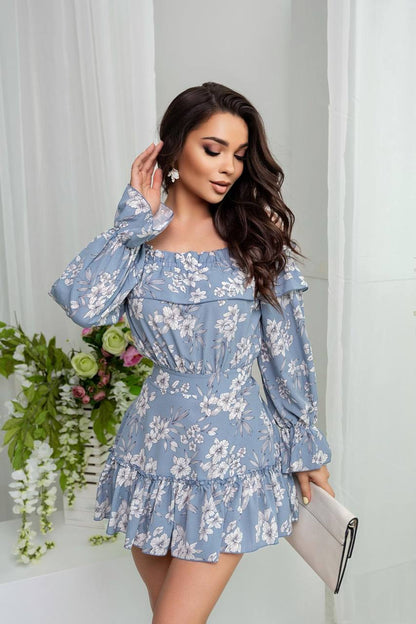 Woman wearing a blue floral dress holding a clutch against a white curtain background