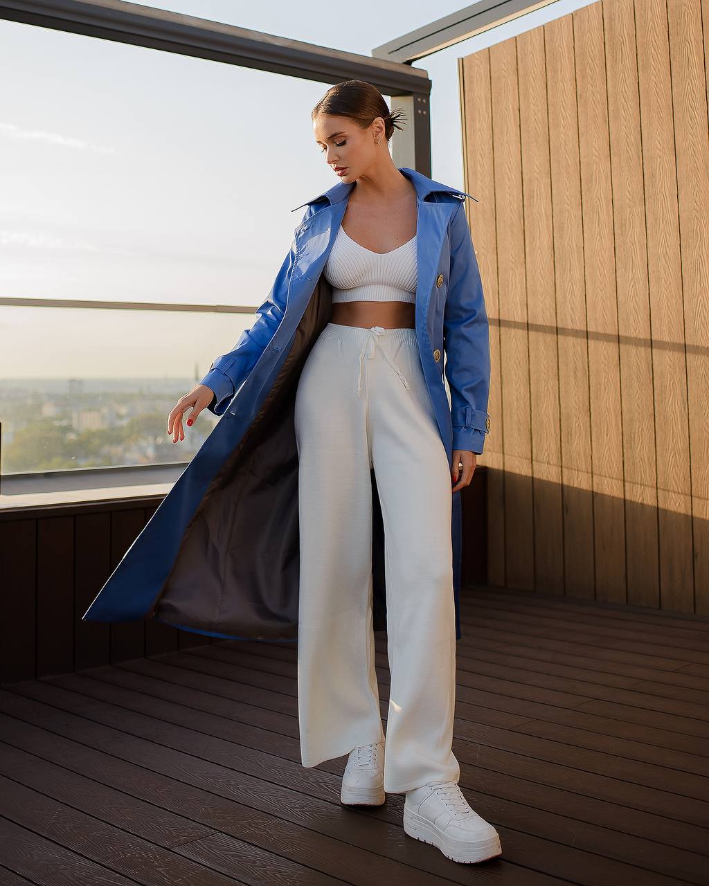 Woman wearing a blue coat and white pants on a wooden deck with a scenic view.