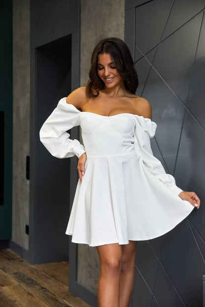 Woman wearing a white off-shoulder dress against a dark tiled wall.