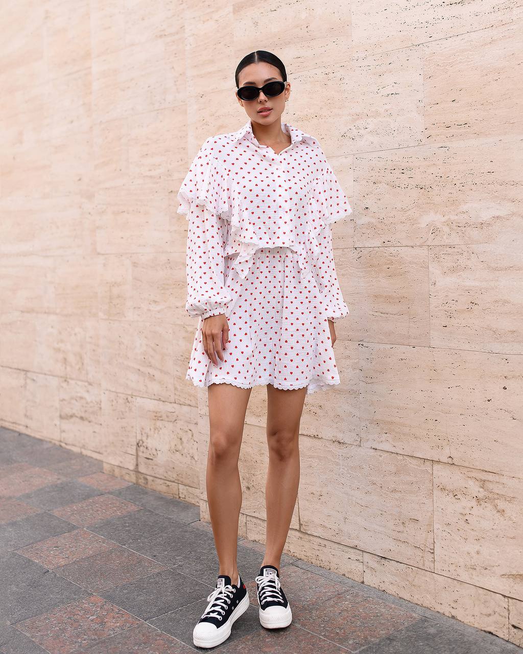 Woman wearing a white polka dot dress with black sneakers against a beige wall.