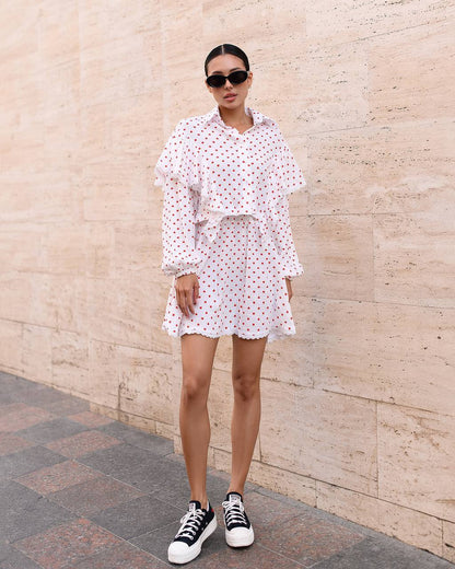 Woman wearing a white polka dot dress with black sneakers against a beige wall.