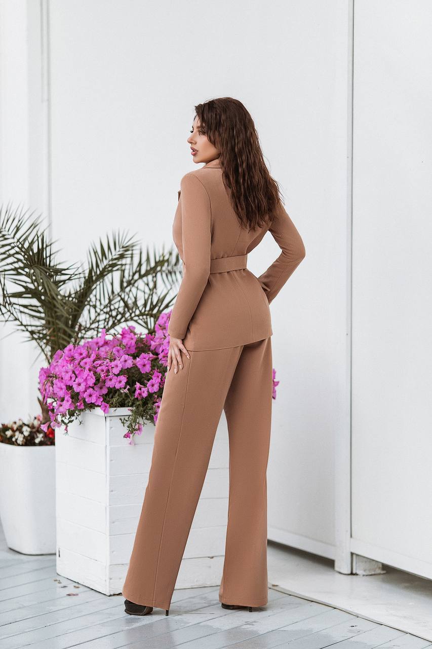 Woman in a brown jumpsuit standing in a room with white walls and potted plants.