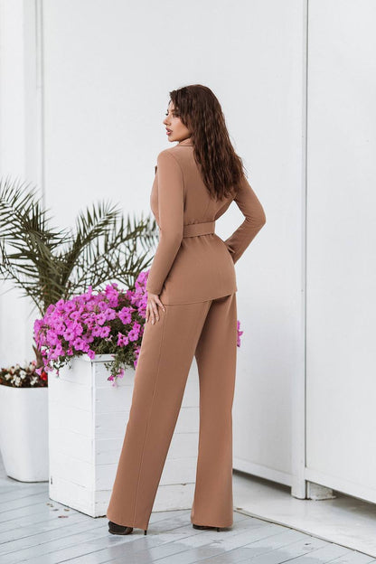 Woman in a brown jumpsuit standing in a room with white walls and potted plants.