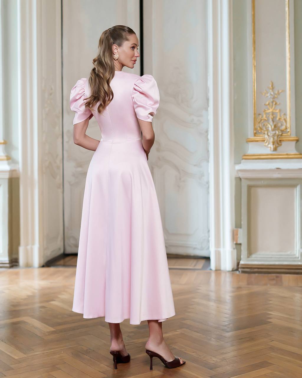 Woman in a pink dress standing in an elegant room with wooden flooring and decorative wall.