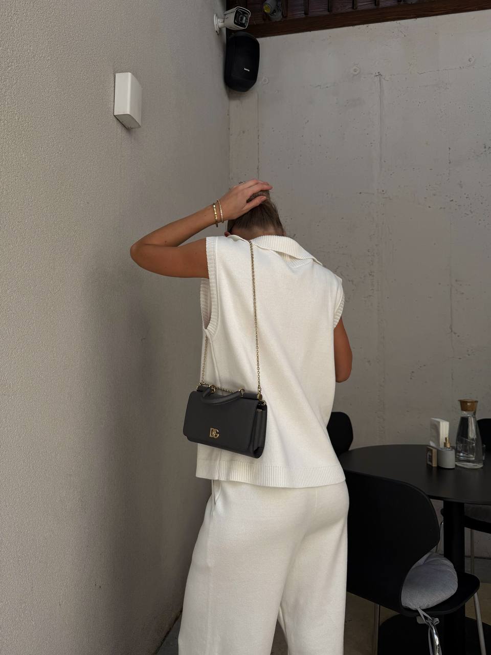 Person in a white outfit with a black handbag standing against a plain wall.