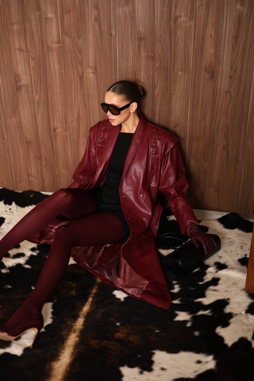 Person wearing a red leather jacket and tights sitting on a cowhide rug against a wooden wall.