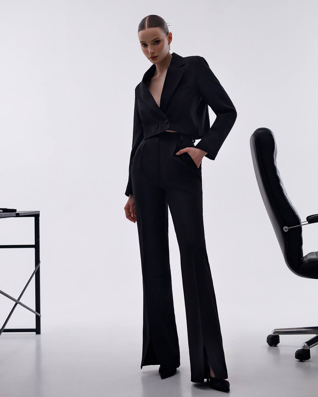 Woman in a black suit standing in a minimalistic office setting with a chair and desk.