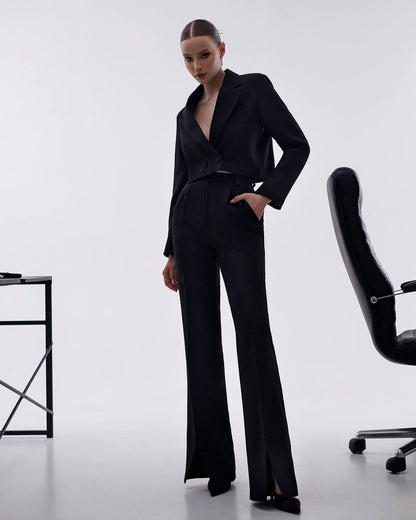 Woman in a black suit standing in a minimalistic office setting with a chair and desk.